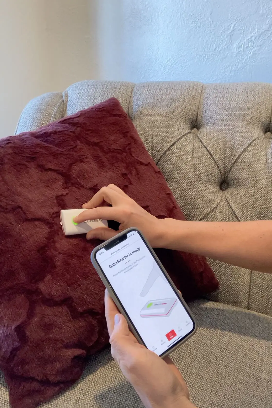 A hand holding a Datacolor ColorReader EZ device against a plush burgundy pillow to scan and match the perfect color.